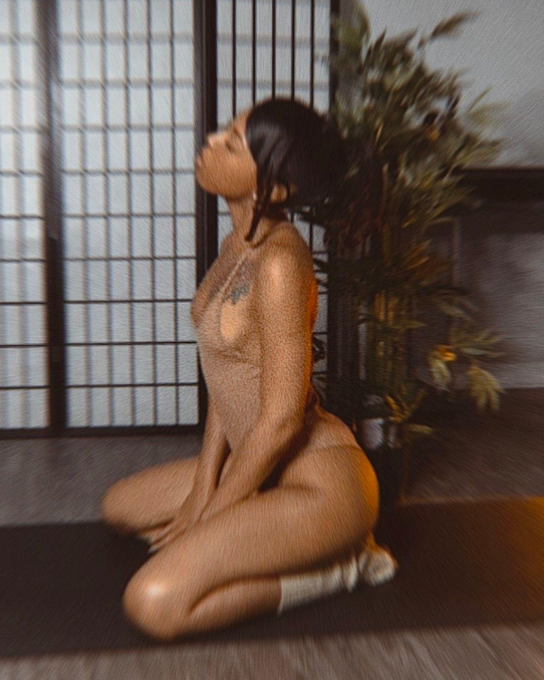 Blurred image of a woman kneeling indoors with a screen and plant in the background.